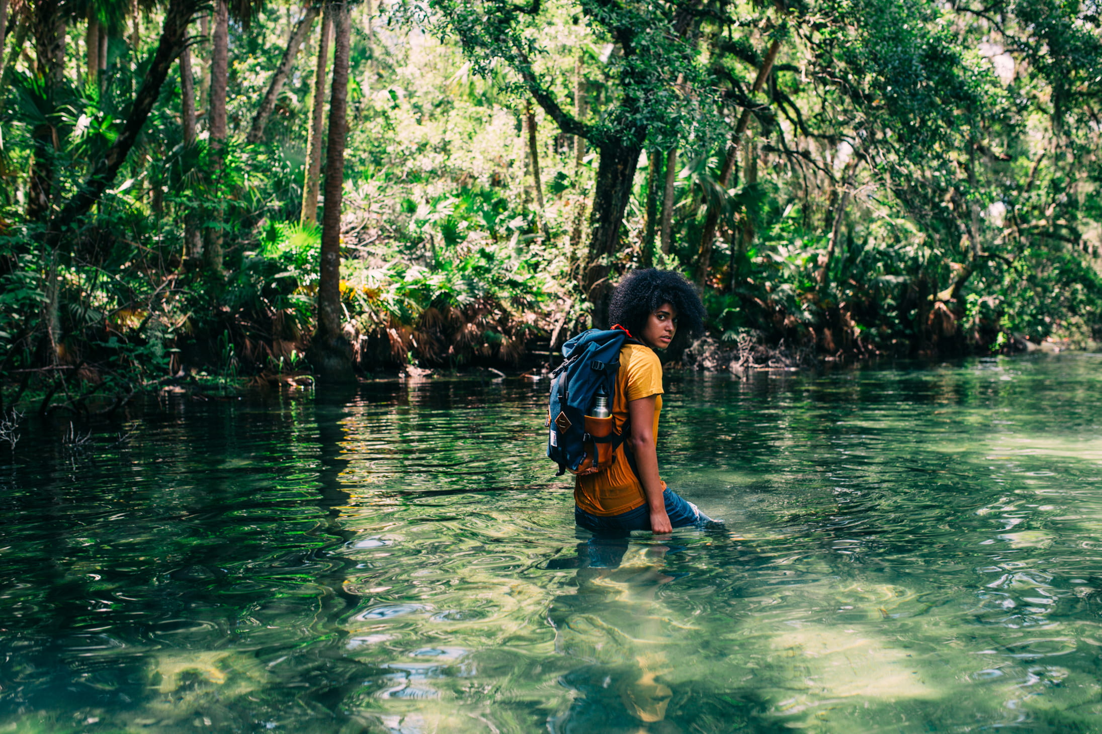 Person in an orange top wearing a backpack walking on a body of water in forest during daytime, man carrying a backpack in a body of water