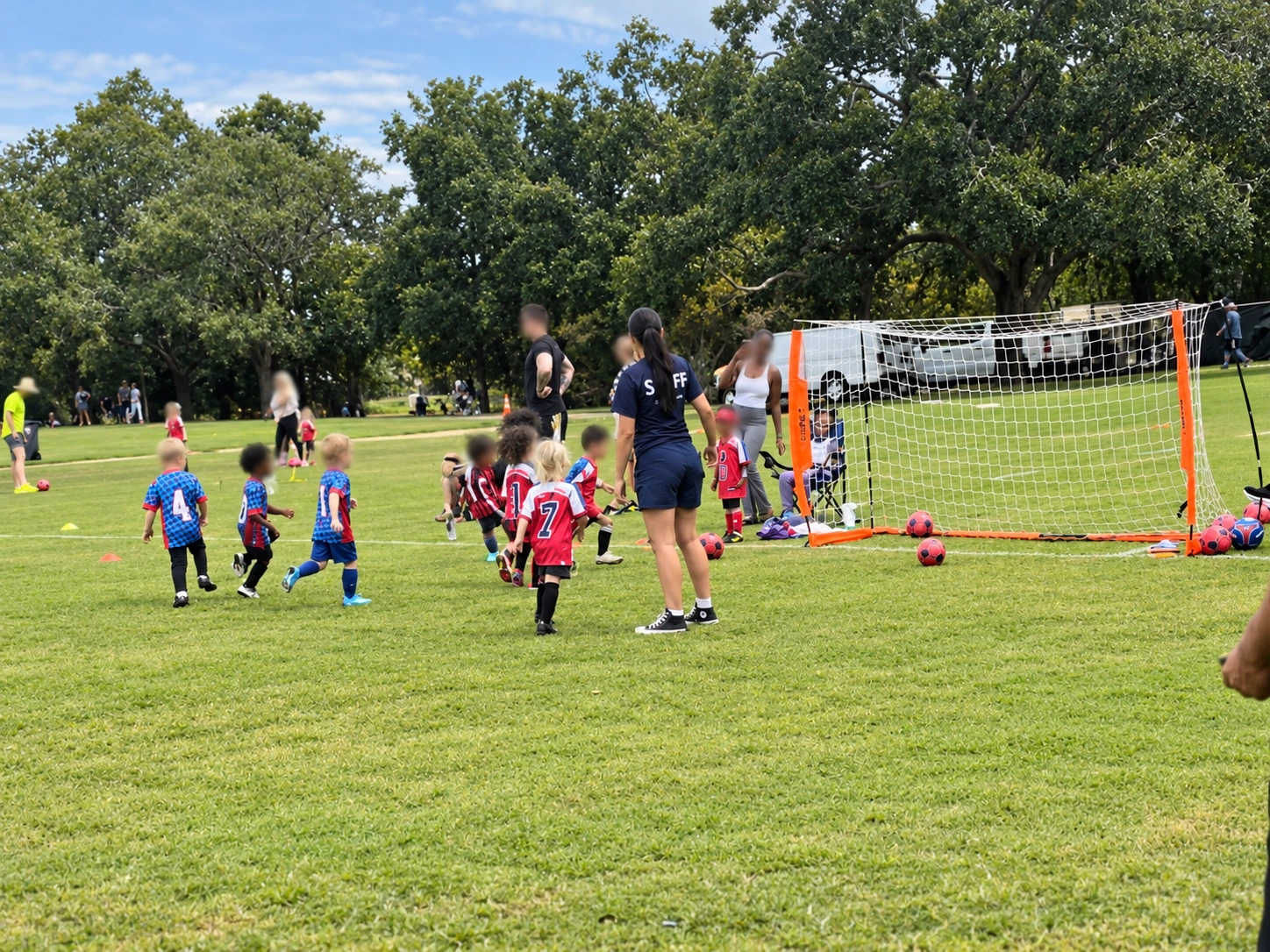 Children in red and blue soccer uniforms play near a portable goal while adults supervise. Participants' faces have been blurred for privacy.