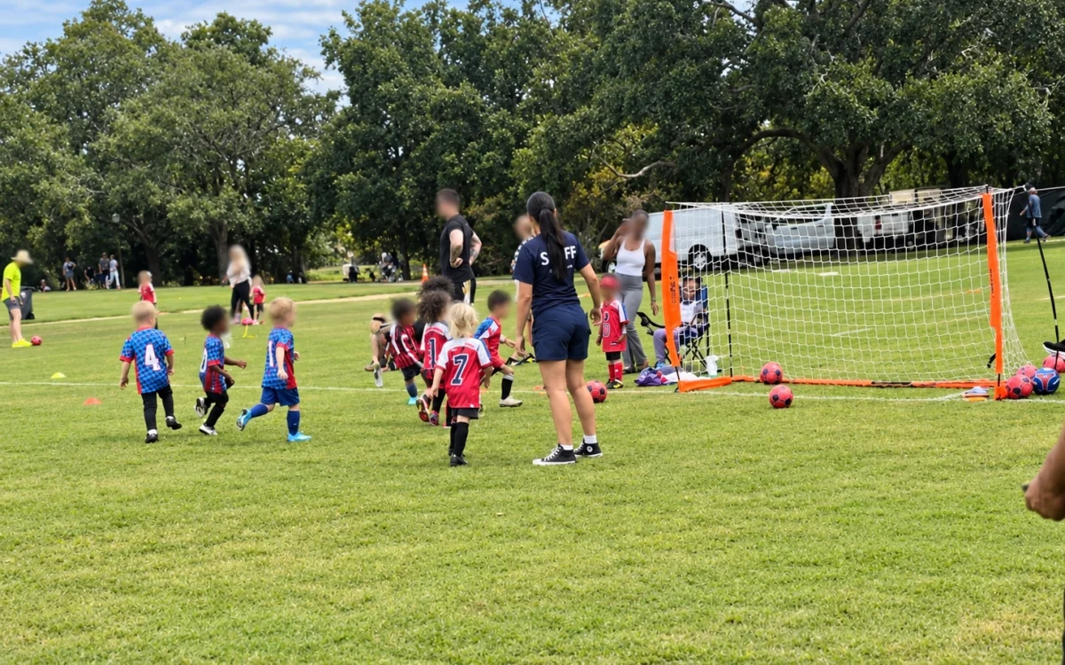 Children in red and blue soccer uniforms play near a portable goal while adults supervise. Participants' faces have been blurred for privacy.